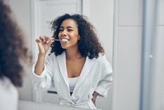 Woman in white robe brushing teeth in bathroom mirror