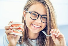 Girl with black framed glasses holding sample braces and Invisalign