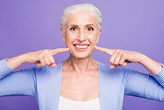 An older woman wearing a purple sweater and pointing to her new smile in McMinnville