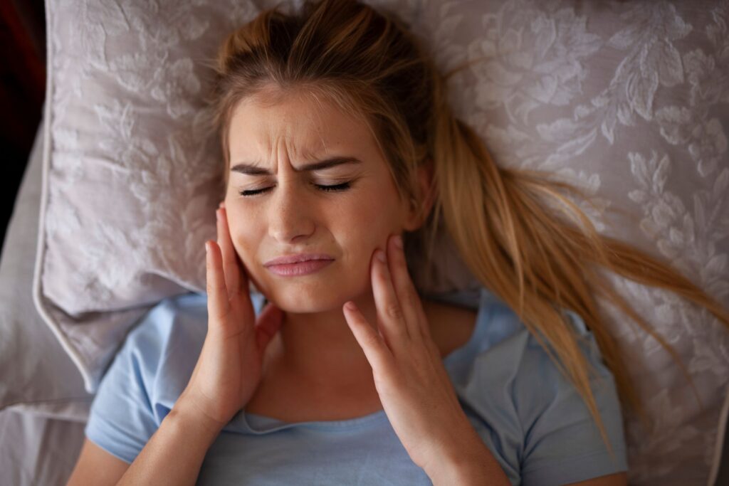 Woman with light brown hair laying on a pillow holding both hands to her aching jaw